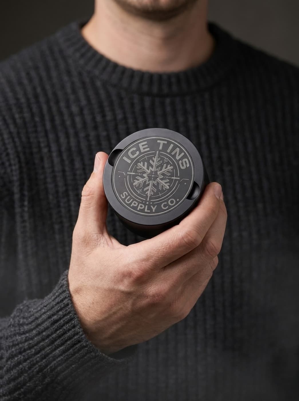 The Ice Tin held in one hand, roughly the width of a palm, against a charcoal knit sweater